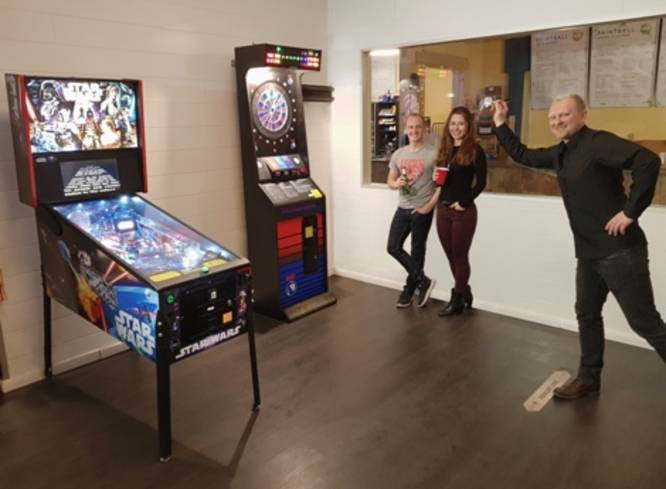 Person spielt Dart, 2 schauen zu. Flipperautomat im Bild.