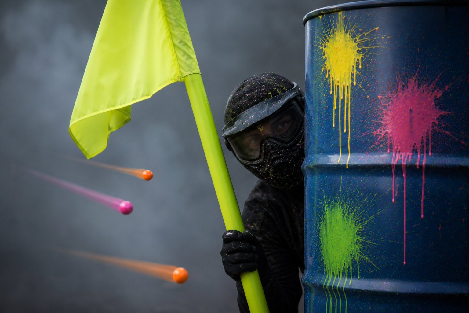 Paintball-Spieler mit Maske hält hinter einem bunten Fass die Flagge, Farbkugeln fliegen vorbei